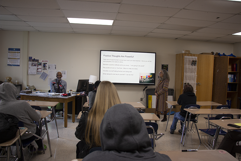 A woman stands in the front of a classroom in front of a screen that advises the students to think positive.