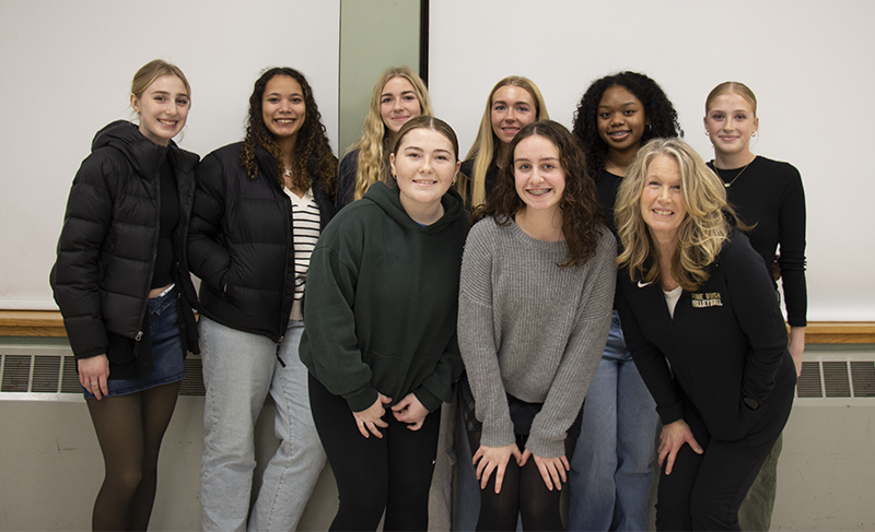 A group of seven high school girls and a woman stand in two rows. They are all smiling.