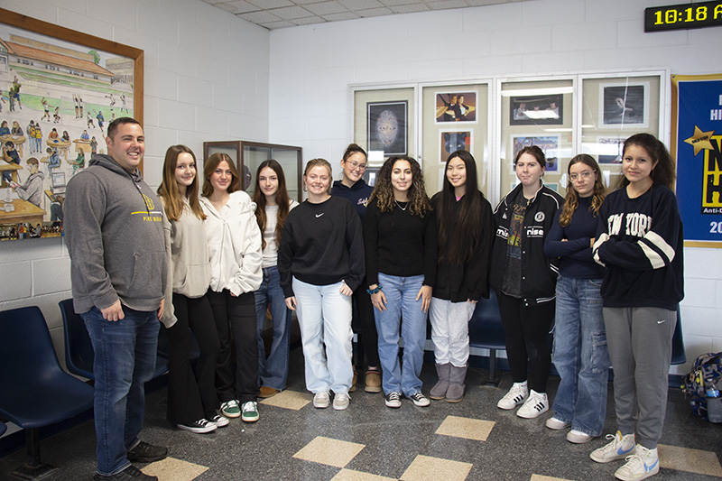 A group of 10 high school girls and a man in a gray sweatshirt stand together smiling.