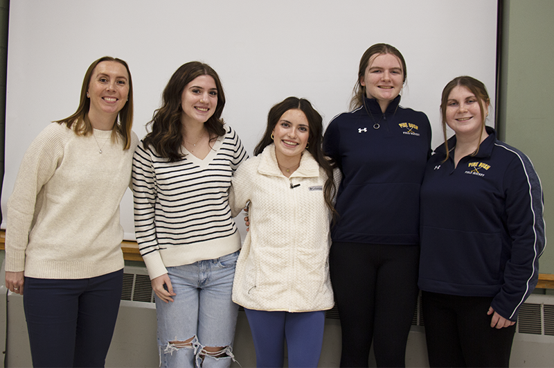 Four young women and a woman who is their coach, stand together arm in arm, for a picture. They are all smiling.