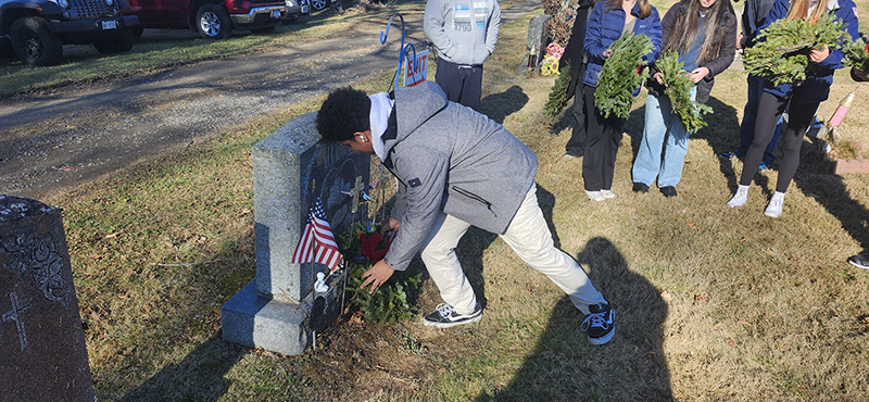 A high school boy bends down to place an evergreen wreath at a gravesite.