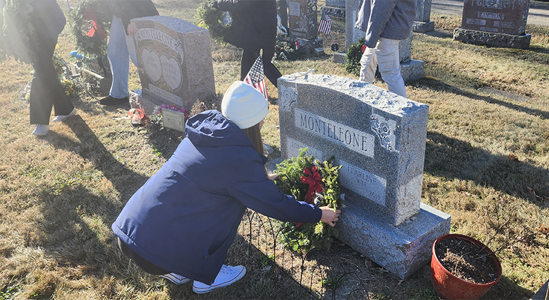 A high school girl wearing a white knit hat and a navy blue jacket places an evergreen wreath at a gravesite.