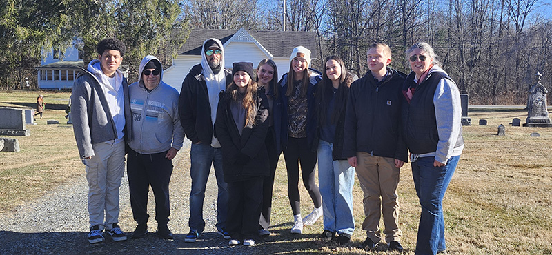 A group of three adults and six high school students stand together They are in a cemetery and dressed for winter.