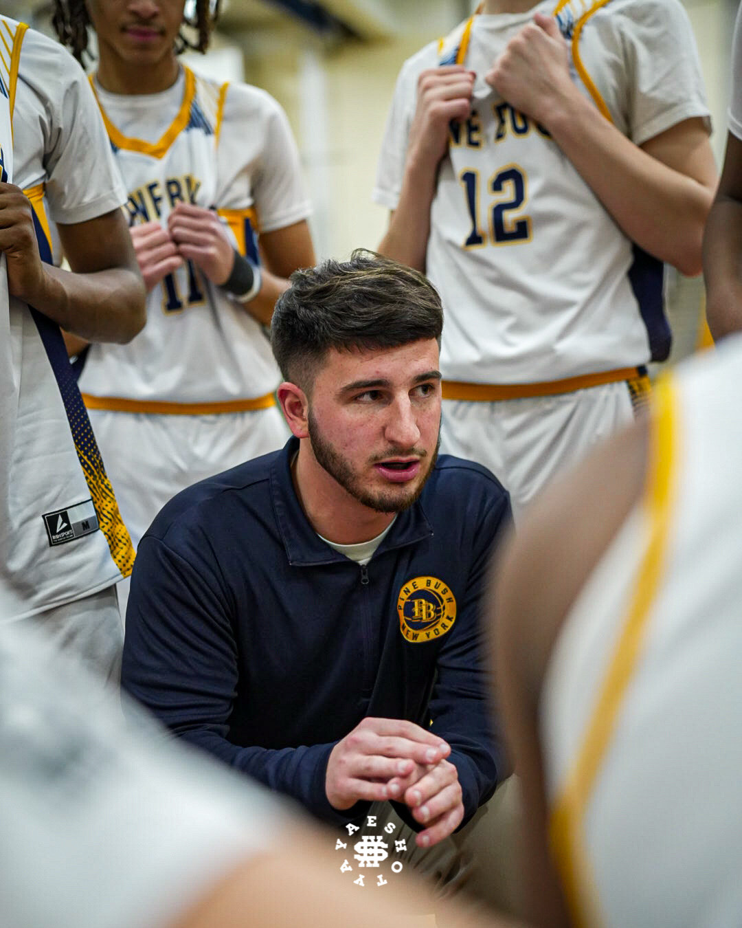 A man with short dark hair and a close cut beard squats down in the middle of a circle of basketball players all wearing white uniforms.