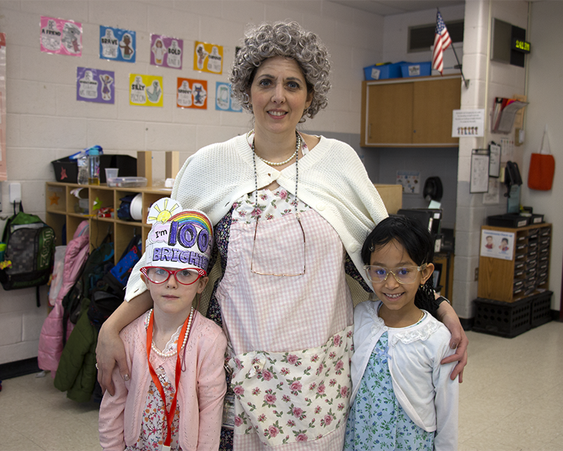 A woman wears a short curly gray wig, glasses on a necklace and a cardigan sweater.. She is standing with two little kids dressed as old people.