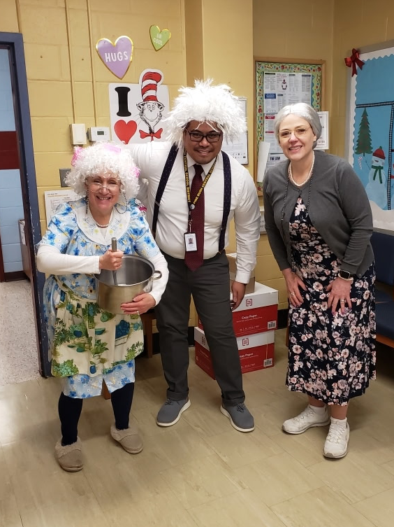 Three adults dress as 100-year-old people. The two women are wearing dresses and sneakers. On ehas a gray wig and the other a white one. The man in the center is wearing a white wig.