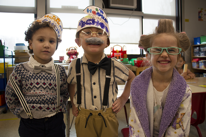 Three young elementary students dressed as old people. The little girl has blue glasses and her hair in curlers. The boy in center is wearing suspenders, a bushy mustache and a bow tie. and the oy on the left is wearing a sweater vest. both boys are wearing paper hats that say 100 days.