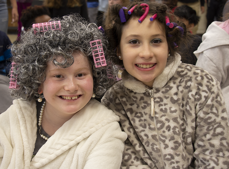 Two older elementary age girls smile. They are dressed as old people with gray hair and curlers in them.