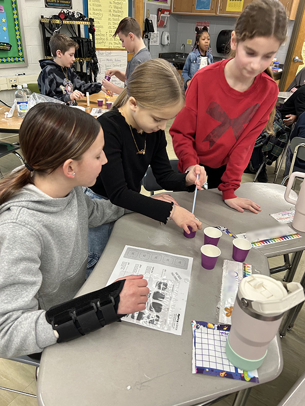 Three fifth-grade girls work together, with cups on their desk and directions for a lab.