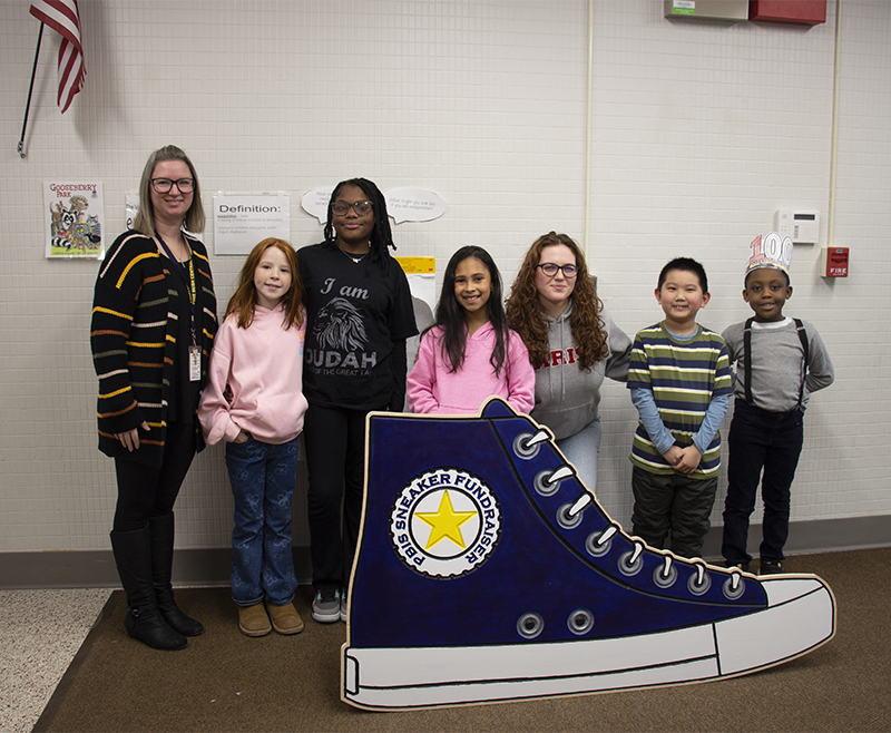 An adult woman stands with five elementary age students and a high school student. They are all smiling. In front of them is a very large wooden cutout of a blue high top sneaker. At the top is a gold star and around it says PBIS Sneaker Fundraiser.