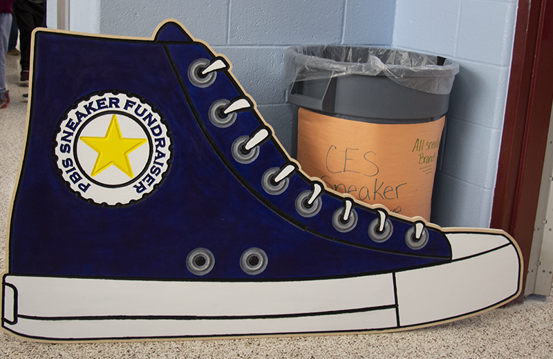 A very large wooden cutout of a blue high top sneaker. At the top is a gold star and around it says PBIS Sneaker Fundraiser. Behind it is a bin that says  CES Sneaker Fundraiser.