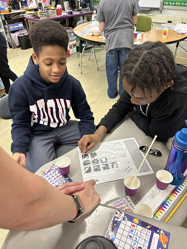 Two fifth-grade boys work together. Their desk is covered in cups and they are reading directions on a piece of paper.