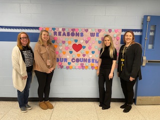 Four woman, two on the left and two on the right, stand by a large poster that says Reasons we Love our counselors. the poster has lots of different colored post-it notes on it.