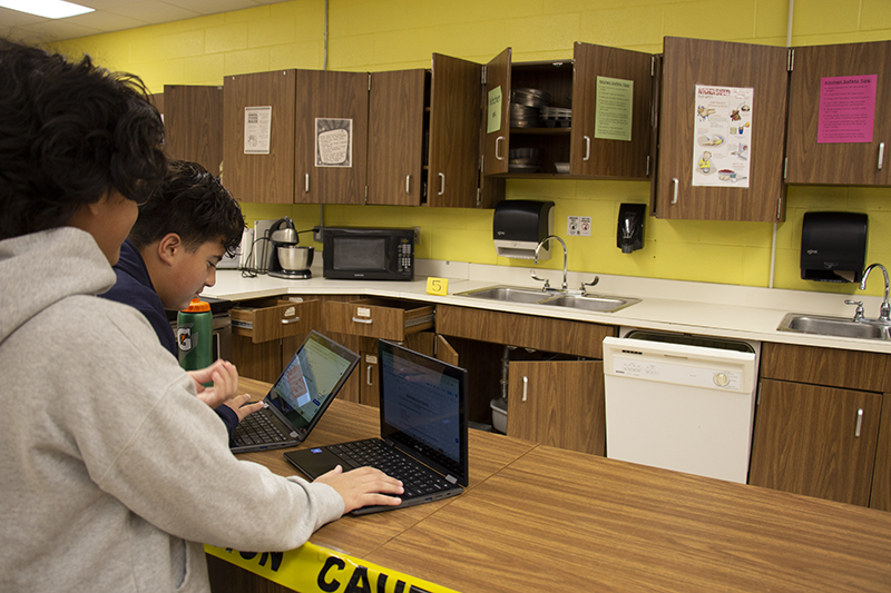 Two 8th-grade boys work on their Chromebooks while looking into a kitchen where the cabinets are all open.