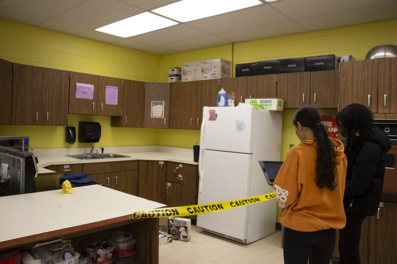 Two 8th-grade girls hold clipboards and look into a kitchen that is marked off with yellow caution tape.