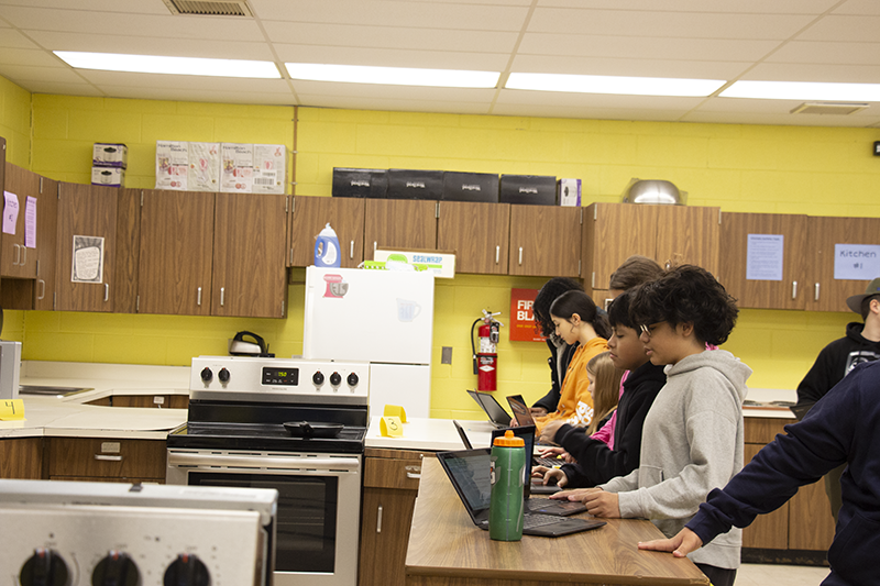 A row of eighth-grade kids stand  working on their Chromebooks, looking into a kitchen.