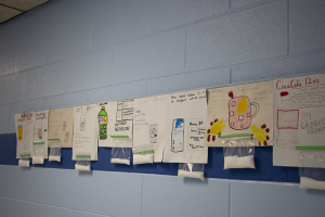 Papers hanging up on a wall, drawn by kids. they show soda, sports drinks and energy drinks with the amount of sugar in each hanging in a bag. 