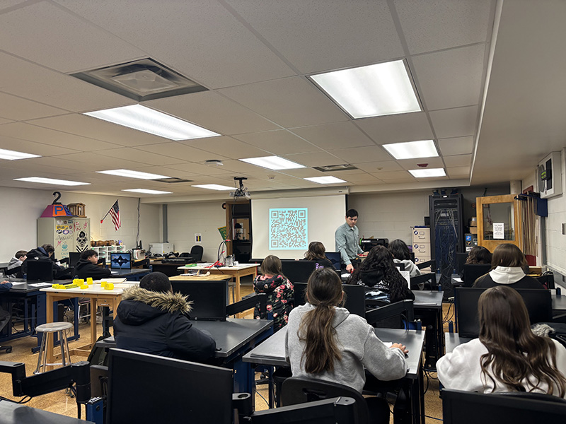 A large classroom with middle school kids sitting at tables listening to a teacher at the front of the room.