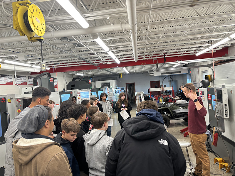 A large group of middle school kids listen while a teacher talks to them. they are in a large classroom where tech is taught.