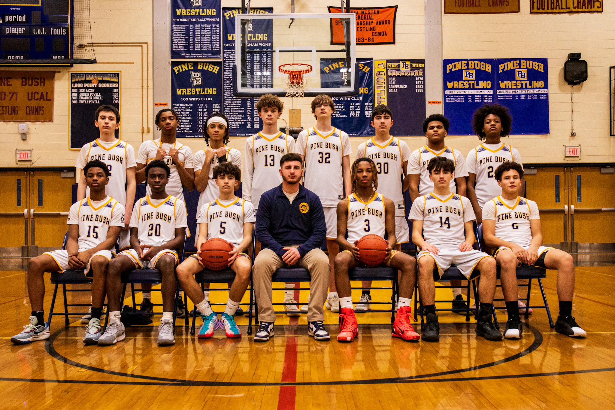 A boys basketball team, i white uniforms, pose for  a team picture. There is a man in a blue jacket sitting in the center.