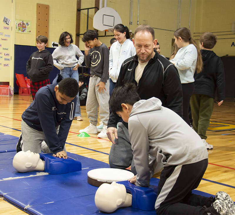 A man watches two fifth-grade boys do CPR to manikins.