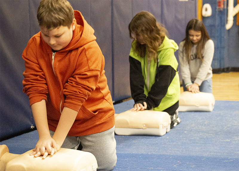 Three fifth-grade kids do CPR on manikins in a line.