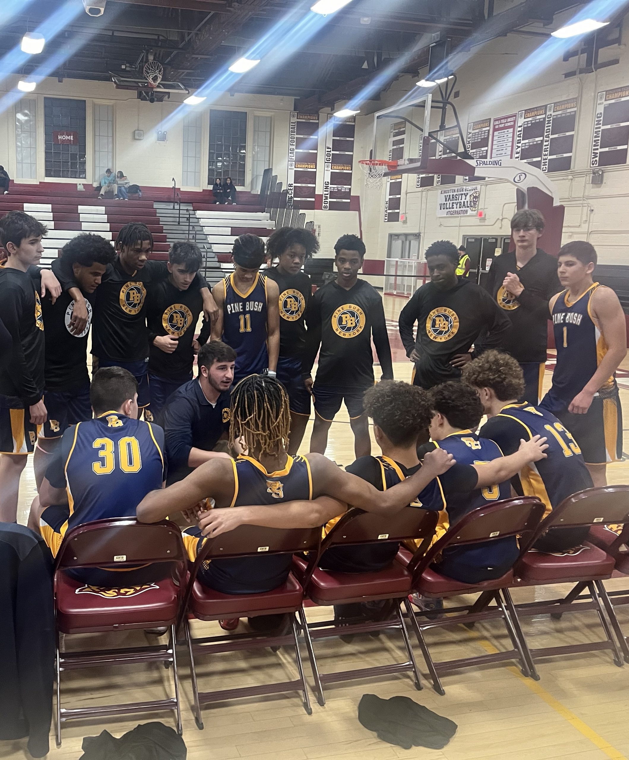 A boys basketball team in navy blue uniforms sit and stand, listening to their coach.