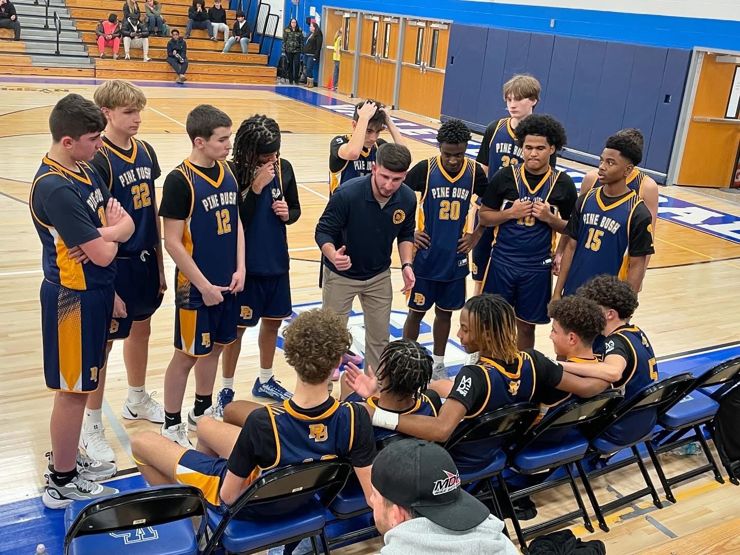 A basketball team in navy blue uniforms  sit in folding chairs and stand in front of them while a man talks to them.