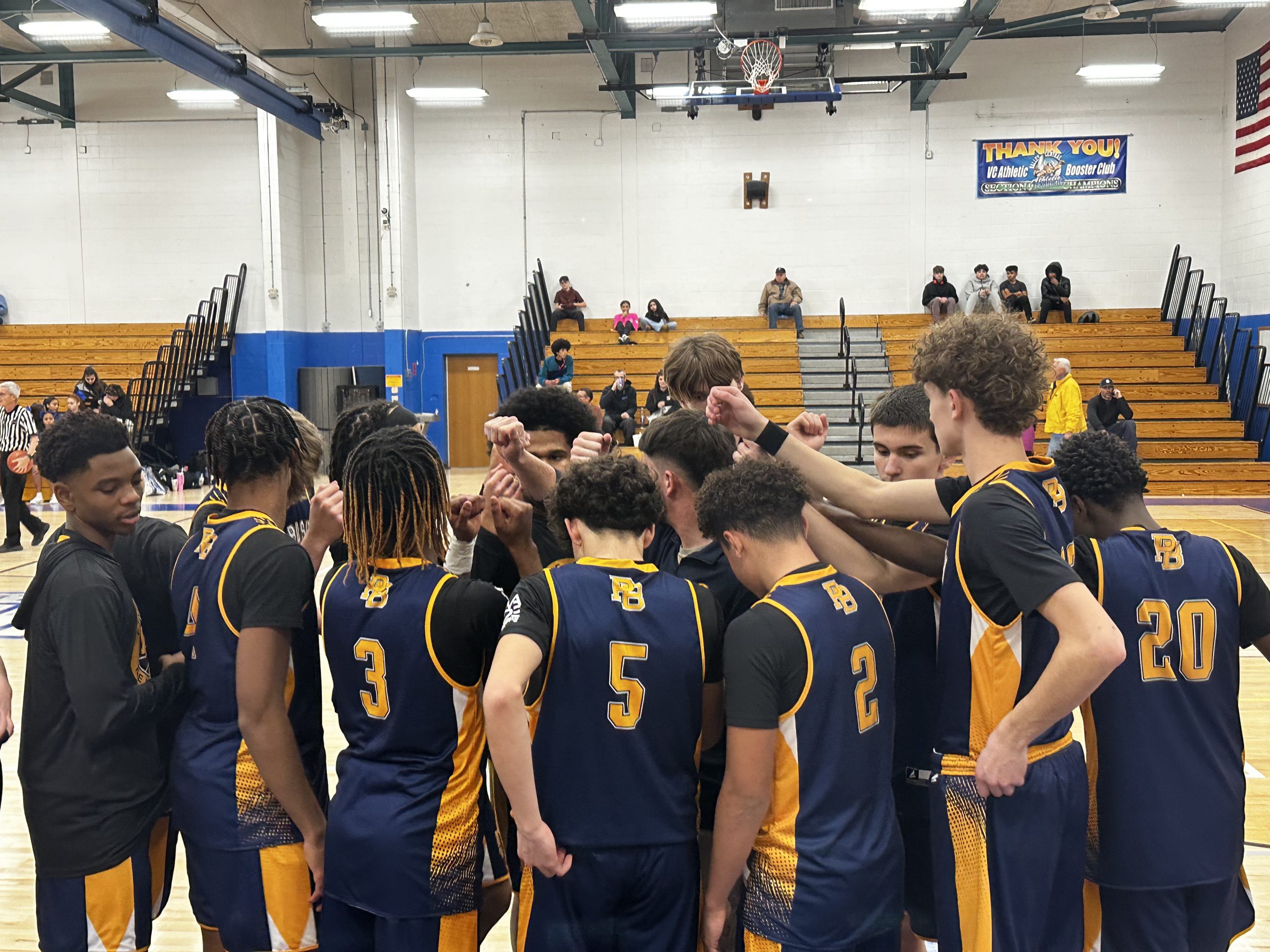 A boys basketball team, in navy blue uniforms stand in a circle with their hands gathered in the middle.