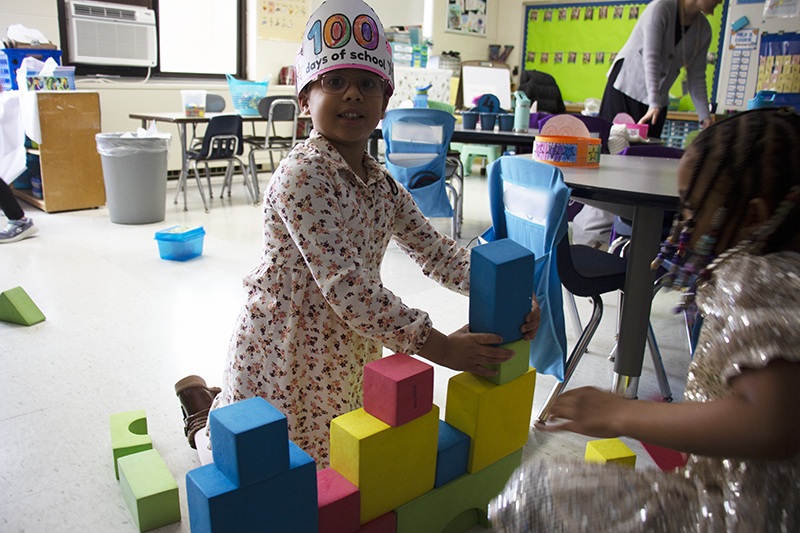 A kindergarten kid wearing a 100 days smarter paper hat builds with multi-color blocks.