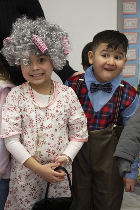 A boy and girl, both kindergarten students, dress as an old couple. The girl has gray hair and curlers and the boy has a bow tie, plaid sweater vest.