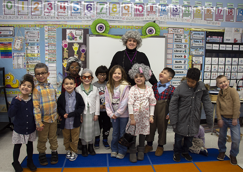 A teacher stands with her class. She is dressed with a gray wig and many of the kids are dressed as old people too.