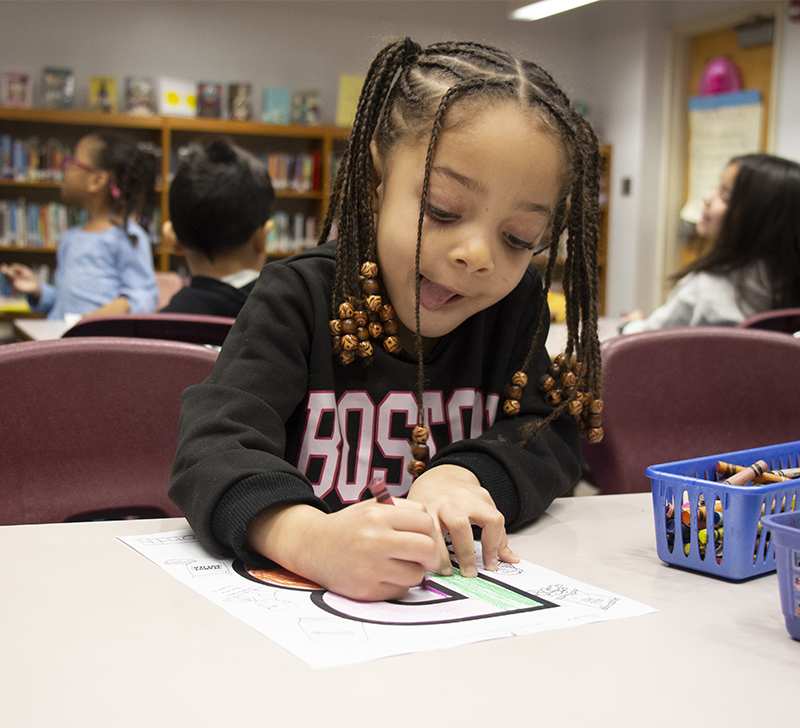 A little girl with braids colors a piece of paper with a big B on it. Her tongue is out a little as she concentrates on her coloring. She is wearing a black shirt with Boston written on it.