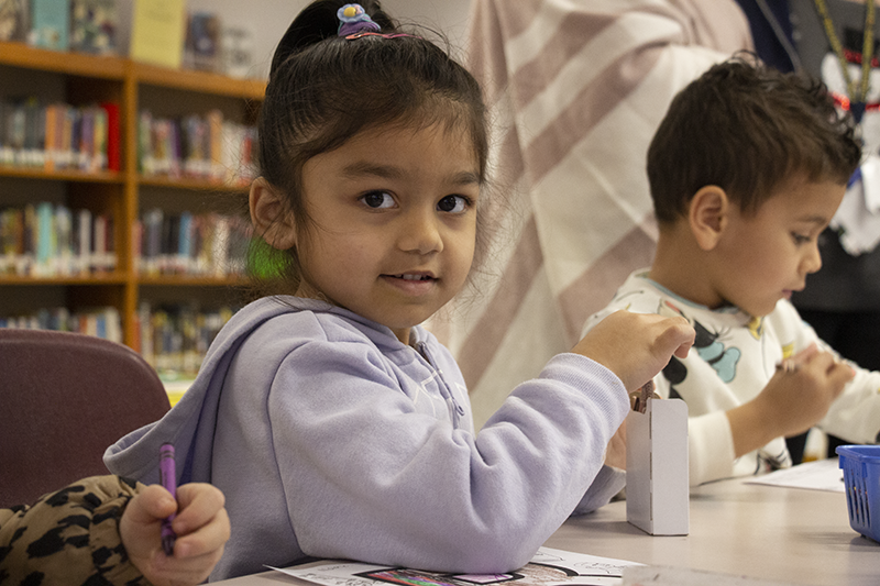 A pre-K girl with a lavender hoodie on smiles at the camera.Her dark hair is pulled up into a ponytail at the top of her head. She is getting a crayon out of a box.