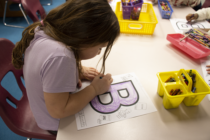 A girl with long dark hair colors a paper with the letter B on it. She is wearing a short-sleeve shirt and looking down at the paper.