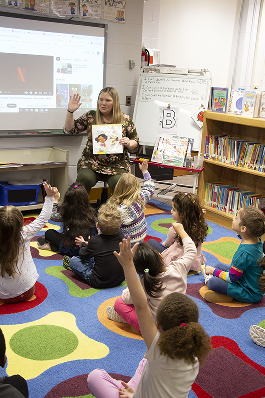 A woman with long blonde hair sits at the front of a class reading from a book she is holding up. A number of pre-K kids sit on the floor listening to her.