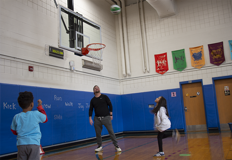 A group of first-grade kids and an adult stand under a basketball basket as one shoots the ball very high.