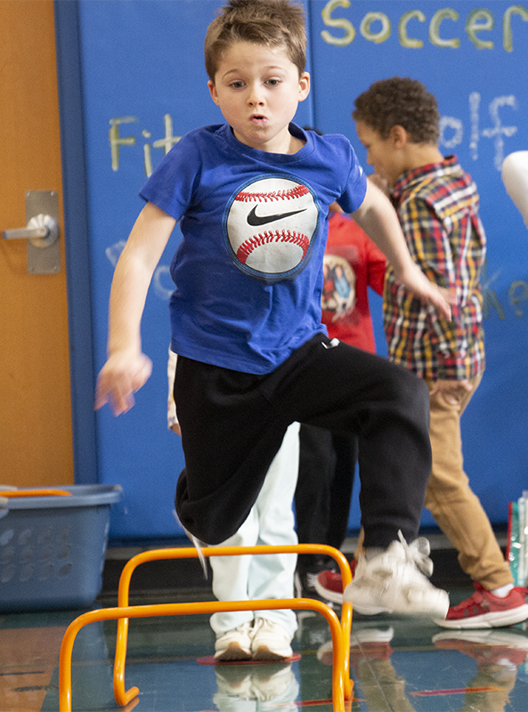 A first-grade boy in a blue shirt with a large baseball on it jumps over an orange hurdle.