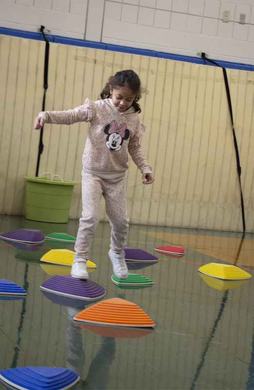 A first-grade girl with a light pink sweatsuit on with Minnie mouse on the shirt, walks from a green stepping stone to a purple one. There are many others around her.