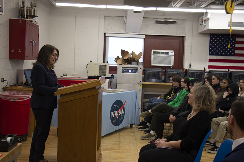 A woman with dark hair wearing a blue pant suit stands at a podium speaking to a room filled with people.