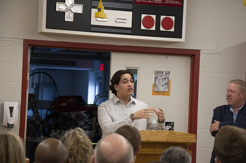 A young man with dark hair stands at a podium and talks to a room filled with people.