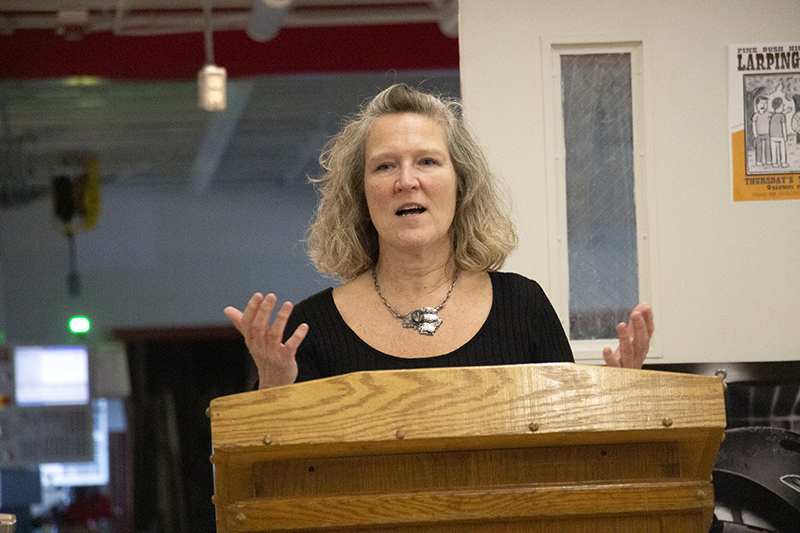 A woman with shoulder-length blonde hair talks at a podium animatedly with her hands.