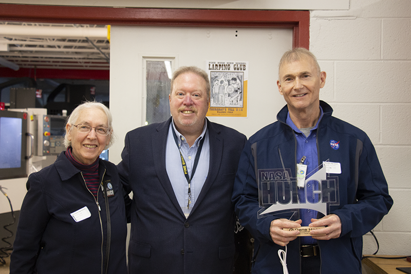 Three adults stand together and smile. A woman on the left, and two men. The man on the right is holding a piece of glass that is etched with the word HUNCH.