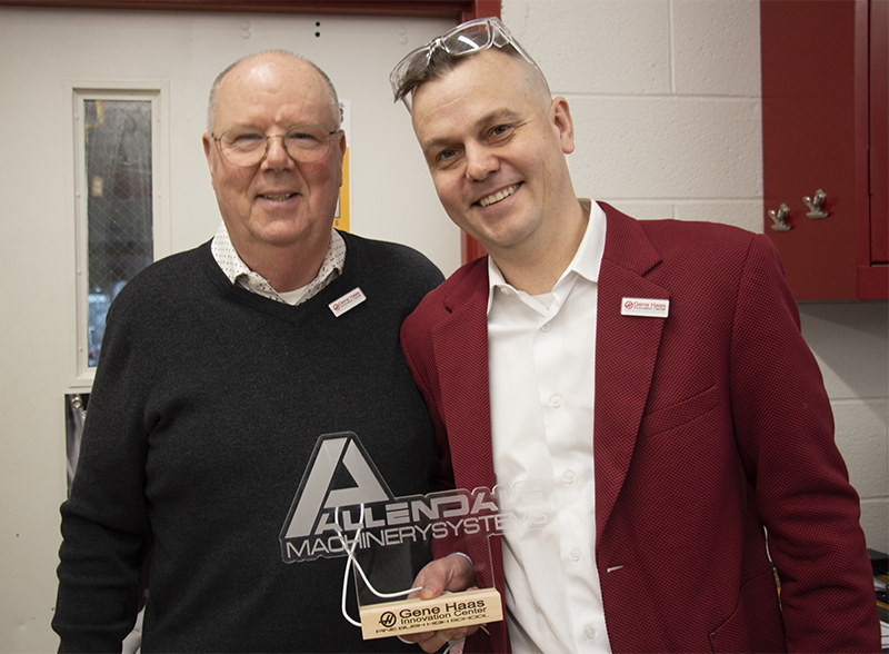 Two men smile. The man on the right is wearing a red suit jacket and white shirt. The man on the left is wearing a white collared shirt and blue sweater. He is holding etched glass that says Allendale Machinery System.