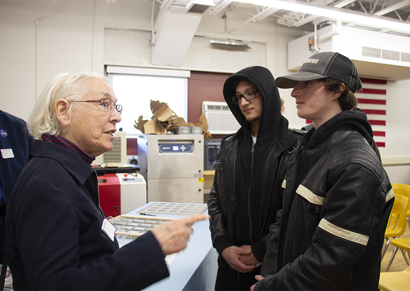 A woman with glasses and white hair talks animatedly to two high school boys.