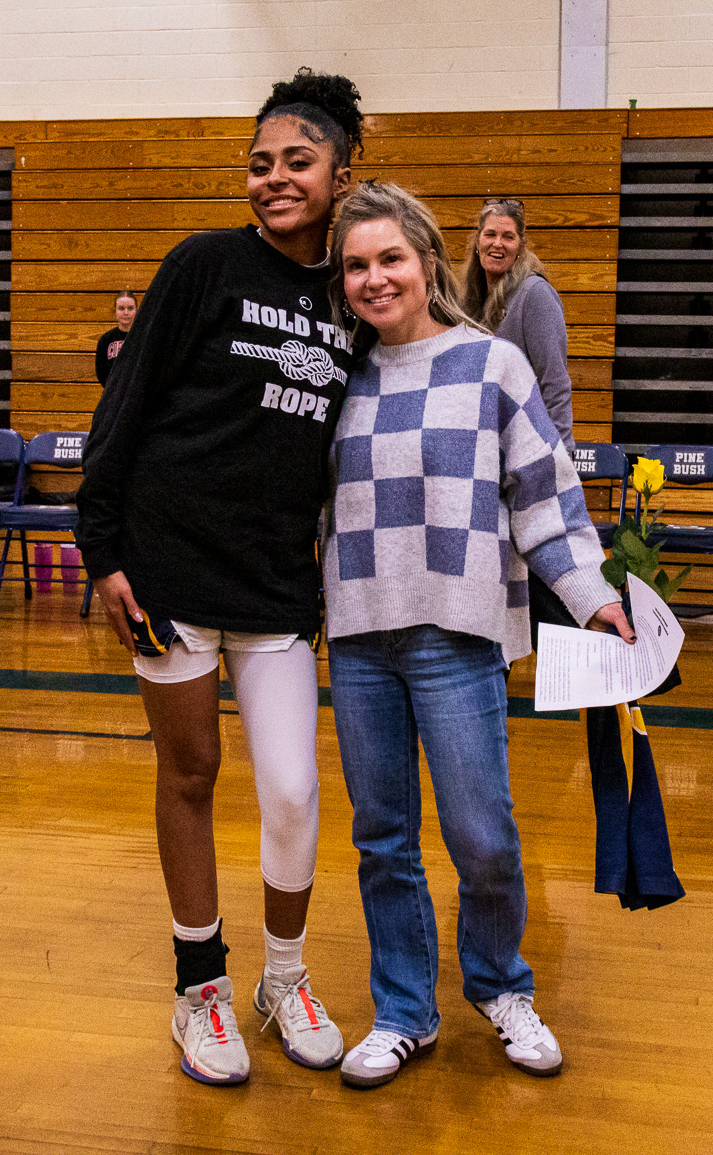 A high school girl has her arm around a woman with long blonde hair, wearing a gray and blue check sweater. They are smiling.