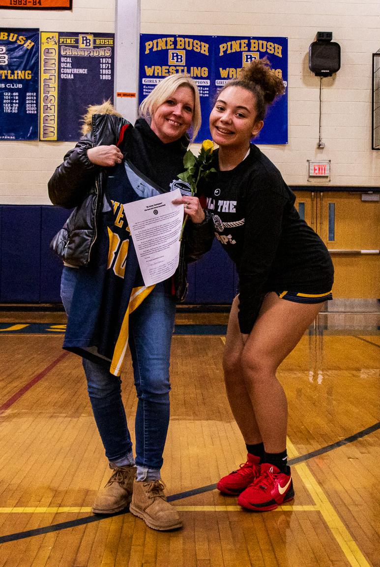 A high school girl on the right has her arm around a woman with blond hair. They are both smiling. The woman on the left is holding a basketball jersey and a flower.