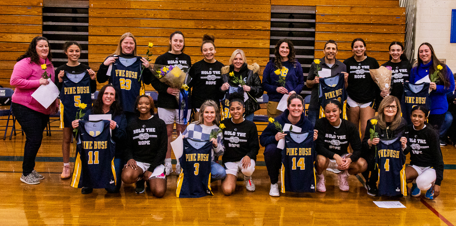 A group of nine high school basketball players  all dressed in blue long-sleeve t-shirts that say Hold the Rope, pose with adults they chose as being instrumental in their lives. Most have flowers and are holding the girls' jerseys.