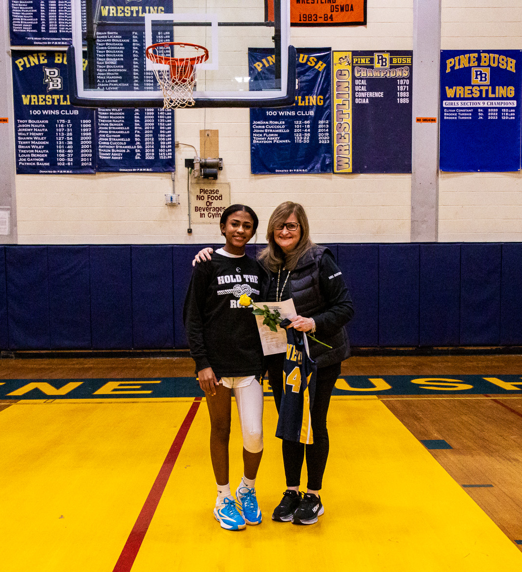 A young woman on the left wearing basketball shorts and a t shirt that says Hold the Rope, has her arm around a woman with long lighter hair. They are both smiling on a basketball court.