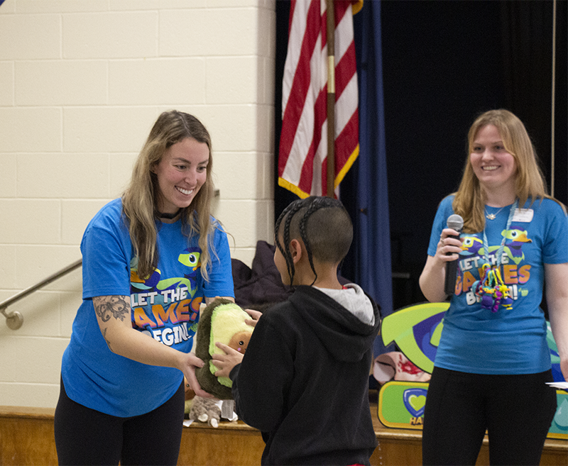 A woman in a bright blue t shirt hands a stuffed avocado to a kid while another woman claps.